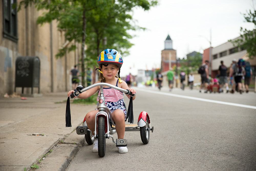 Adorable young girl on a tricycle with a loose helmet looking inquisitive. She hasn't started pedaling yet, and she's parked on the side of the curb.