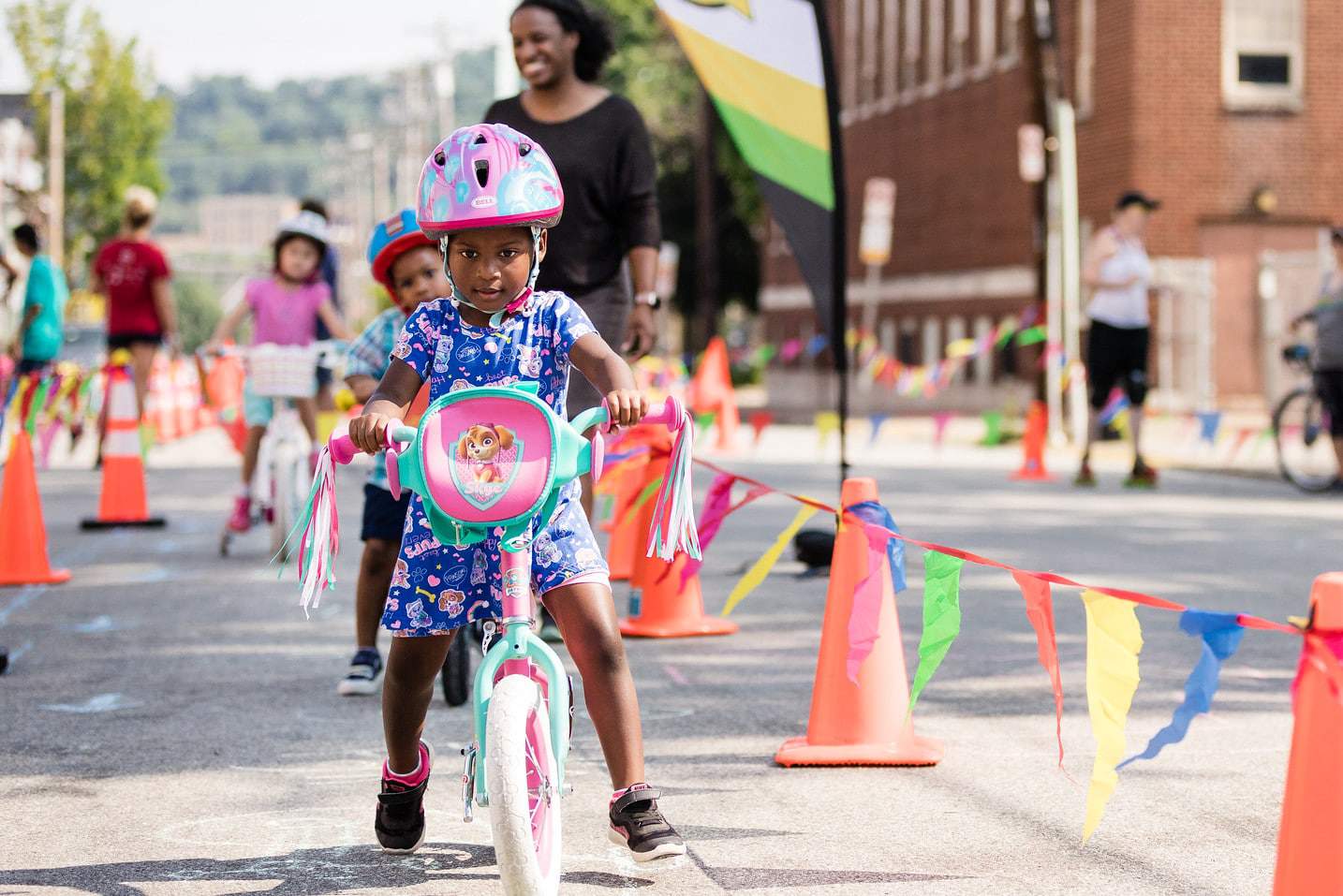 Young girl on a bikes walks her brightly colored bicycle along an obstacle course made of orange cones and flags. She looks focused and adorable! A woman stands behind the kiddo smiling and blurry in the background
