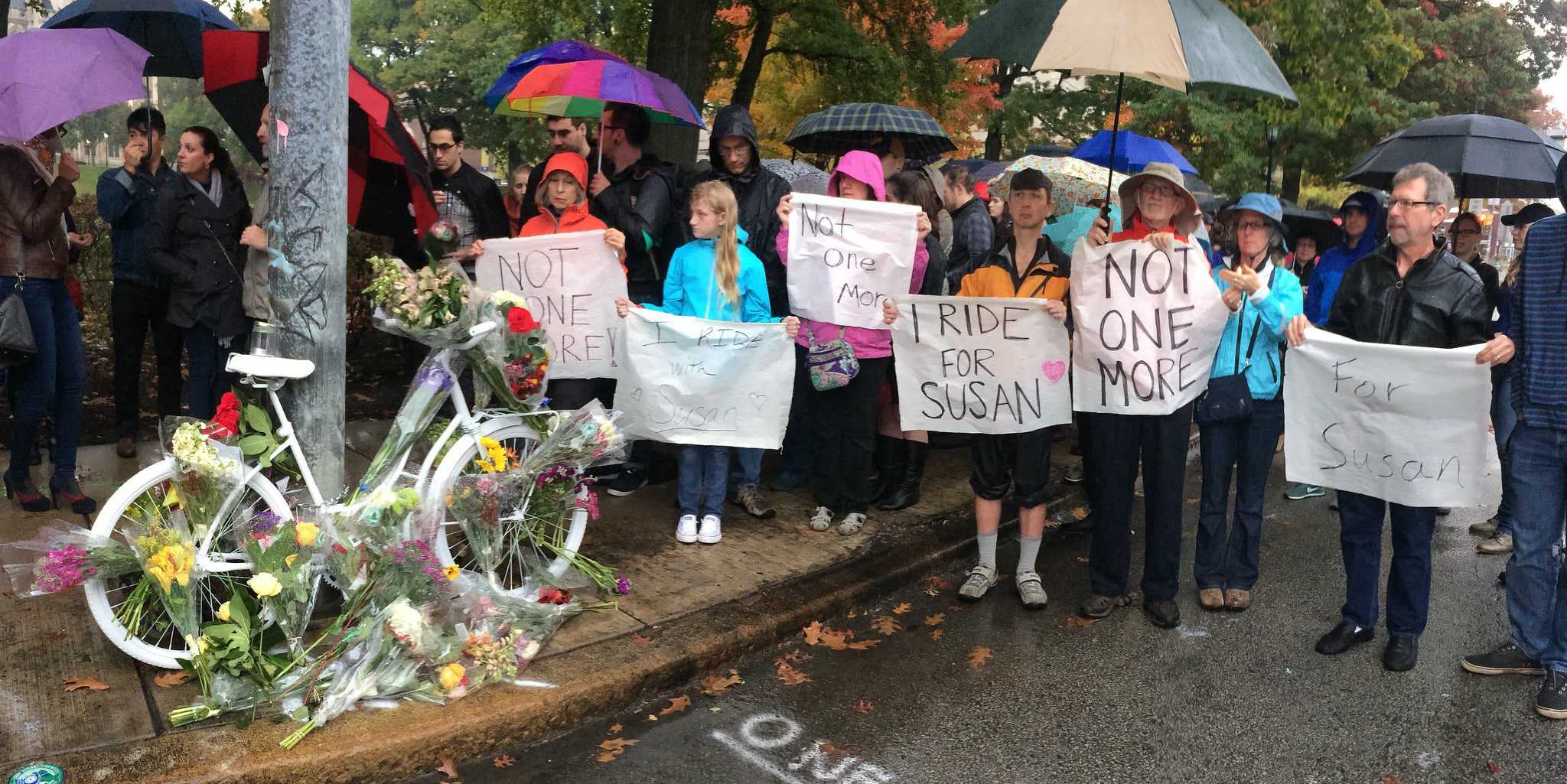 People are gathered around a bike painted white with flower bouquets placed up on it. The people are holding signs that say "not one more" and "I ride for Susan".