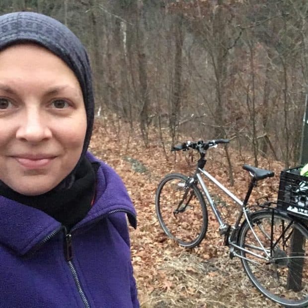 Heather wears a balaklava and sweater. Her bike is in the background with a crate containing extra warm layers.