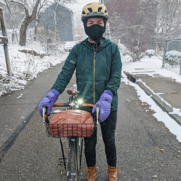 Lydia is standing with her bike in an alleyway. She is wearing a light jacket, mittens, boots, and a face mask.