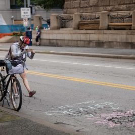 A person stops in the street while biking to take a picture of a pothole with their phone.