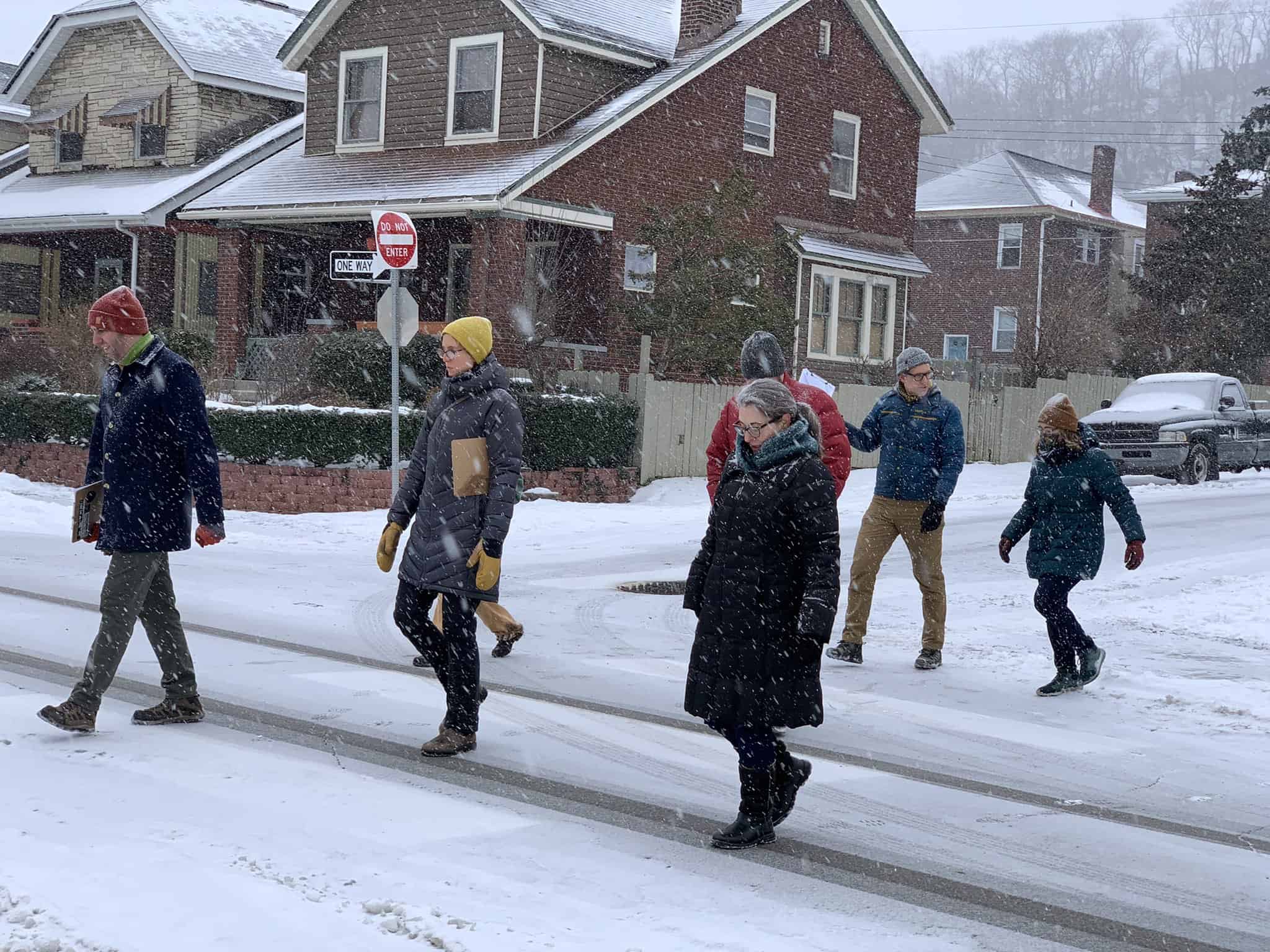 Eight people walk across a snowy intersection holding clipboards.