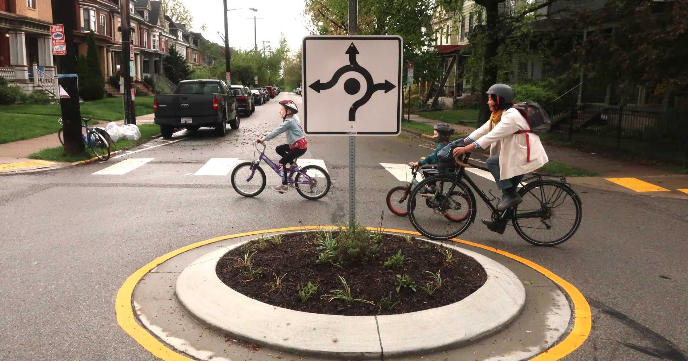 Photograph of two children and an adult woman riding their bikes on a street with a roundabout. 