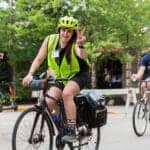 Woman on a bike wearing bright yellow safety vest and bright yellow helmet giving a peace sign with fingers.