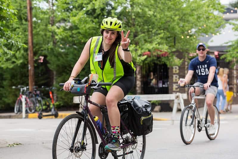 Woman on a bike wearing bright yellow safety vest and bright yellow helmet giving a peace sign with fingers.