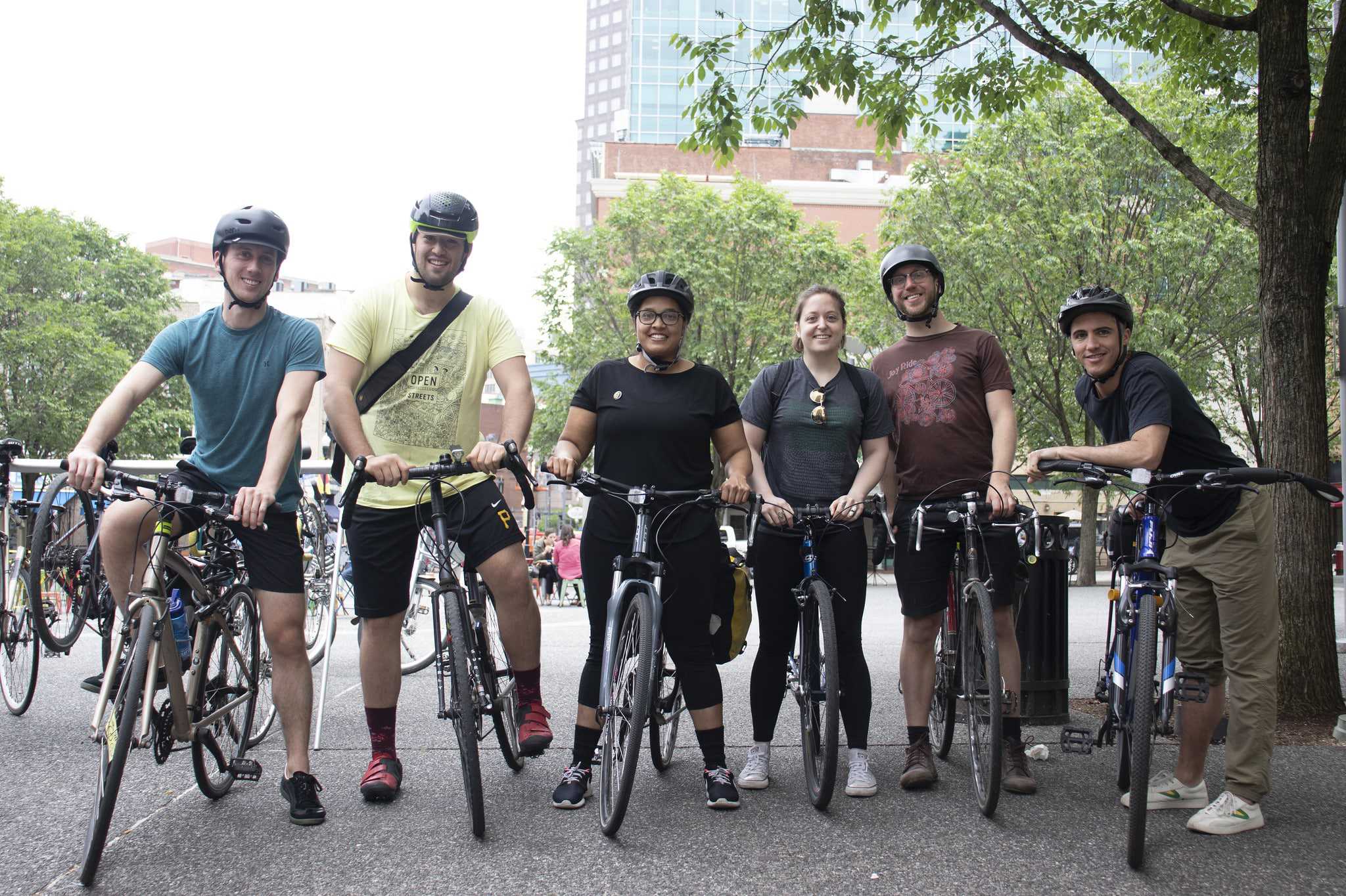 Group of people standing with bikes in a line facing the camera and smiling with city and trees in the background