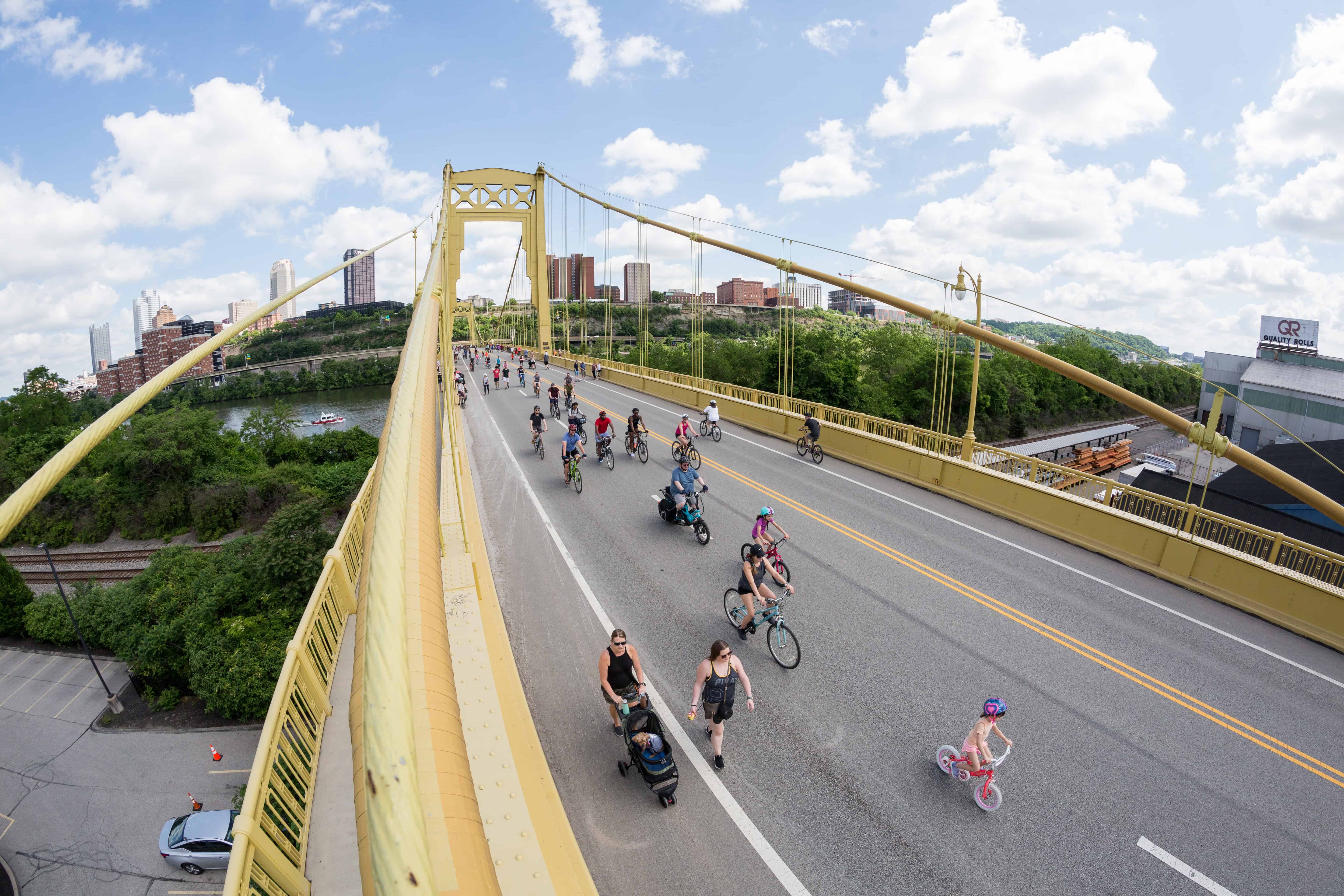 Large yellow suspension bridge with bikers and walkers going across with blue skies and city line in the background.