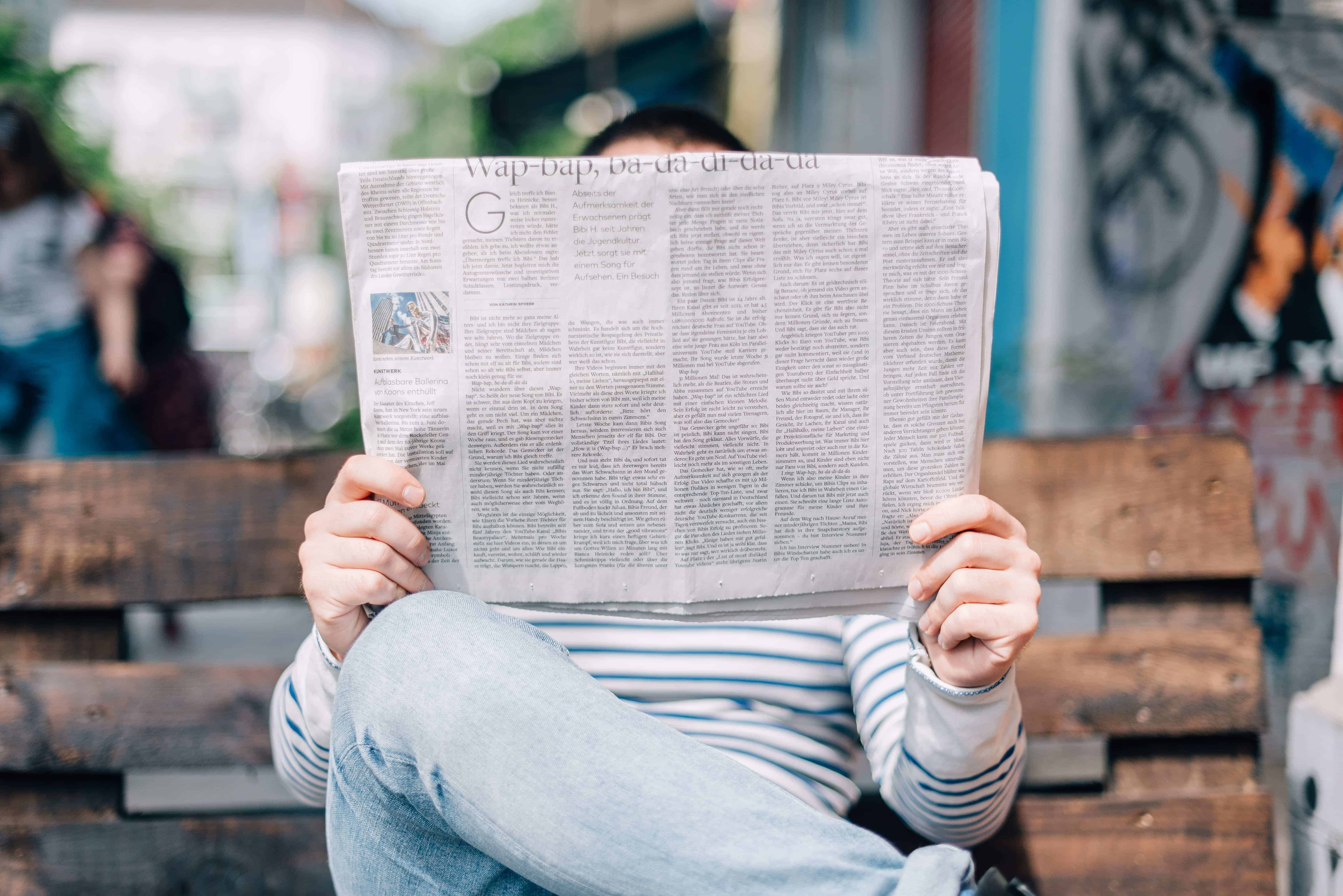 Picture of someone sitting on a bench holding a newspaper in front of their face. The headline is jibberish.