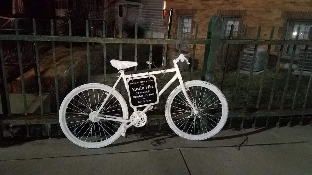 Photo of the Ghost Bike to honor the memory of Austin Fike who was a victim of a traffic crash between a driver and a cyclist.  The bike is painted white with a black plaque that reads "Bicyclist Struck Here. Austin Fike. 22 Years Old. October 20, 2019. Rest in peace."