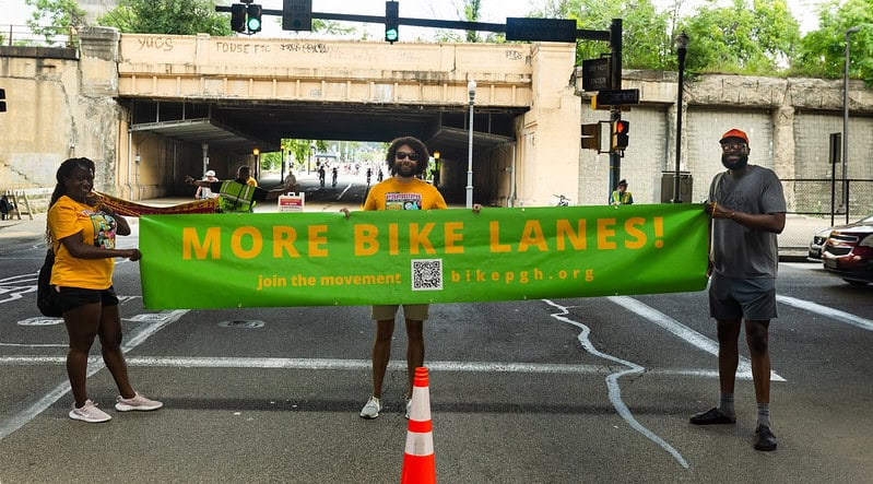 Picture of three people in the middle of the street holding a green banner with yellow text that says "more bike lanes! join the movement bikepgh.org"