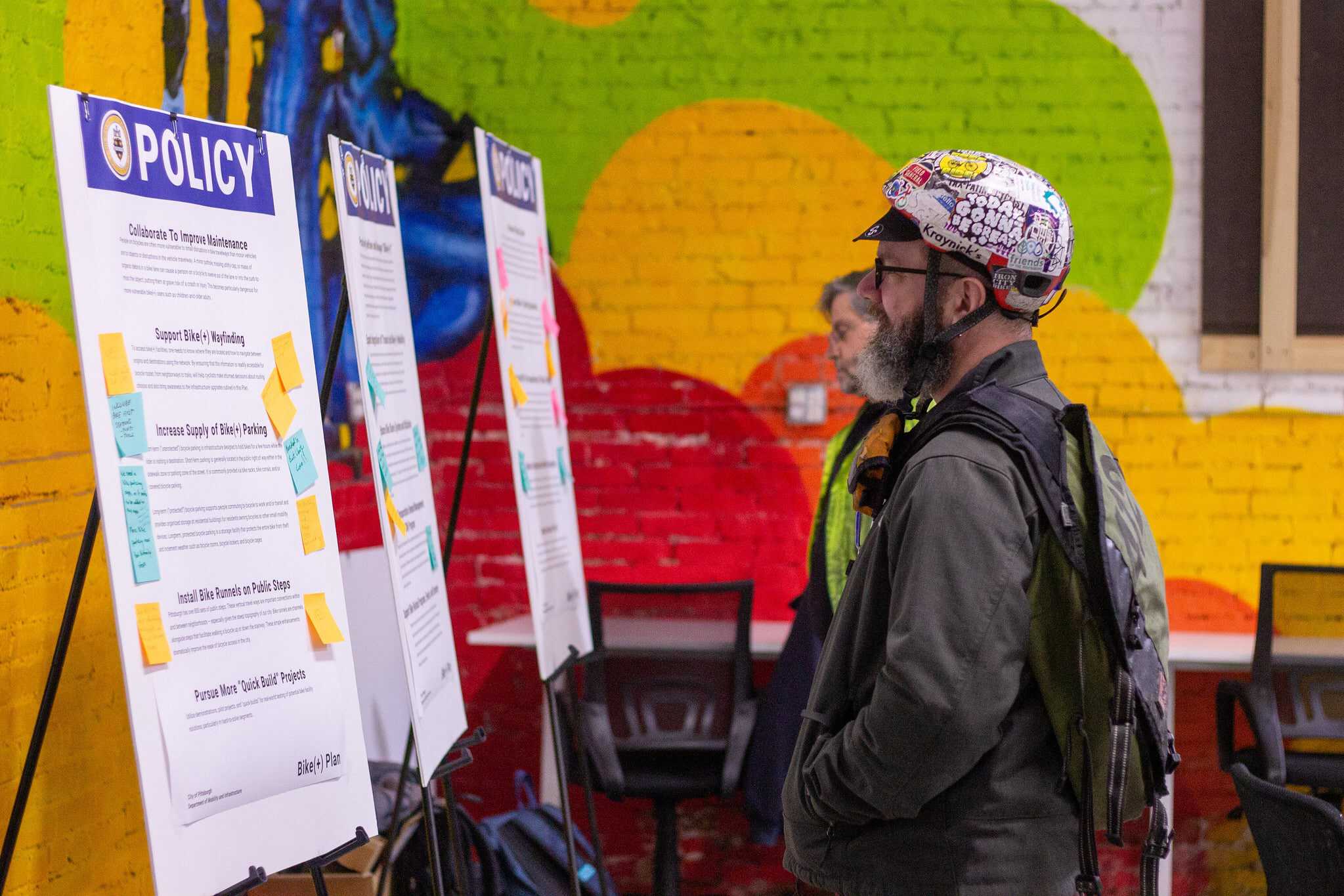 Two people wearing bike gear look at posters at a community meeting that say "policy" at the top in large letters.