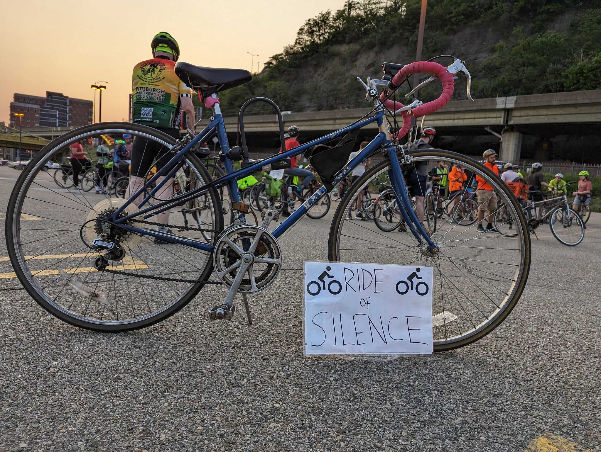 Picture of a blue bike with pink handle bars propped up on a kickstand with a sign leaning on it that says "Ride of Silence" with bike icons on it. The sun is setting in the background with other cyclists in the mid ground.