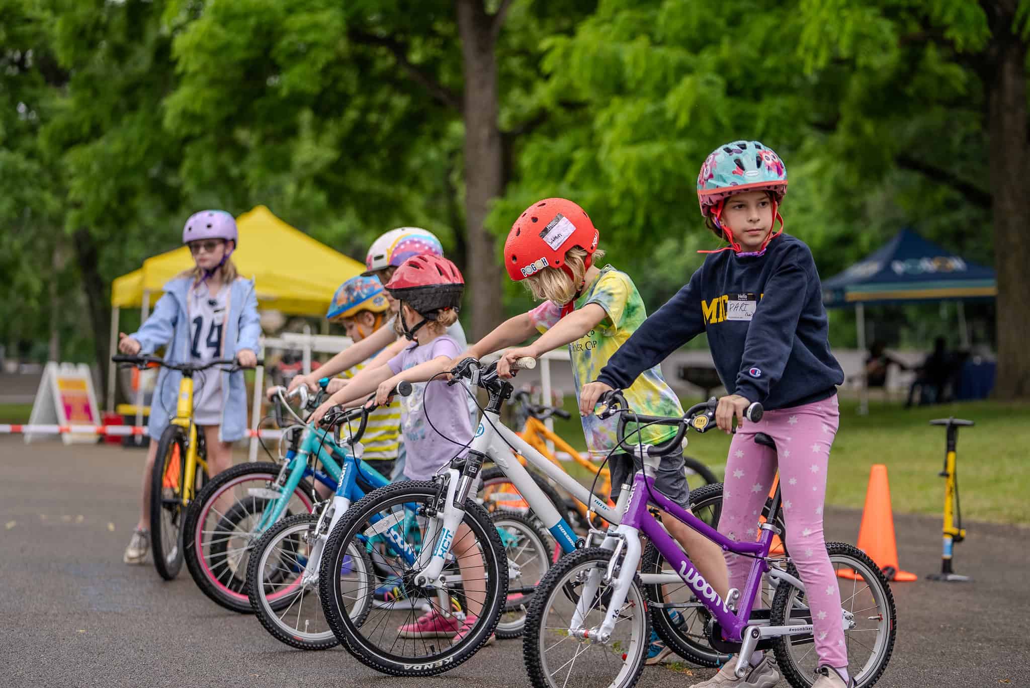 Six children wearing bike helmets and bright clothing stand in a line astride their bicycles. Behind them are tents and cones demarcating a training course.