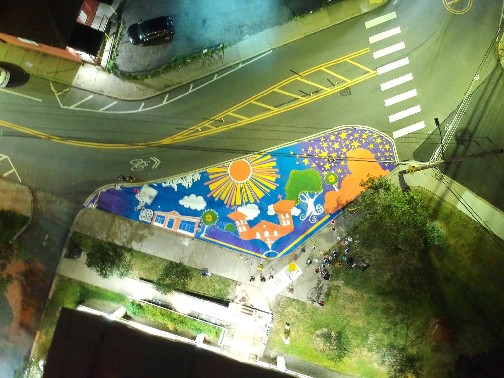 Overhead photo of a street mural featuring the library, a church, and the Allegheny Observatory surrounded by trees with the sun, clouds, and stars above. 