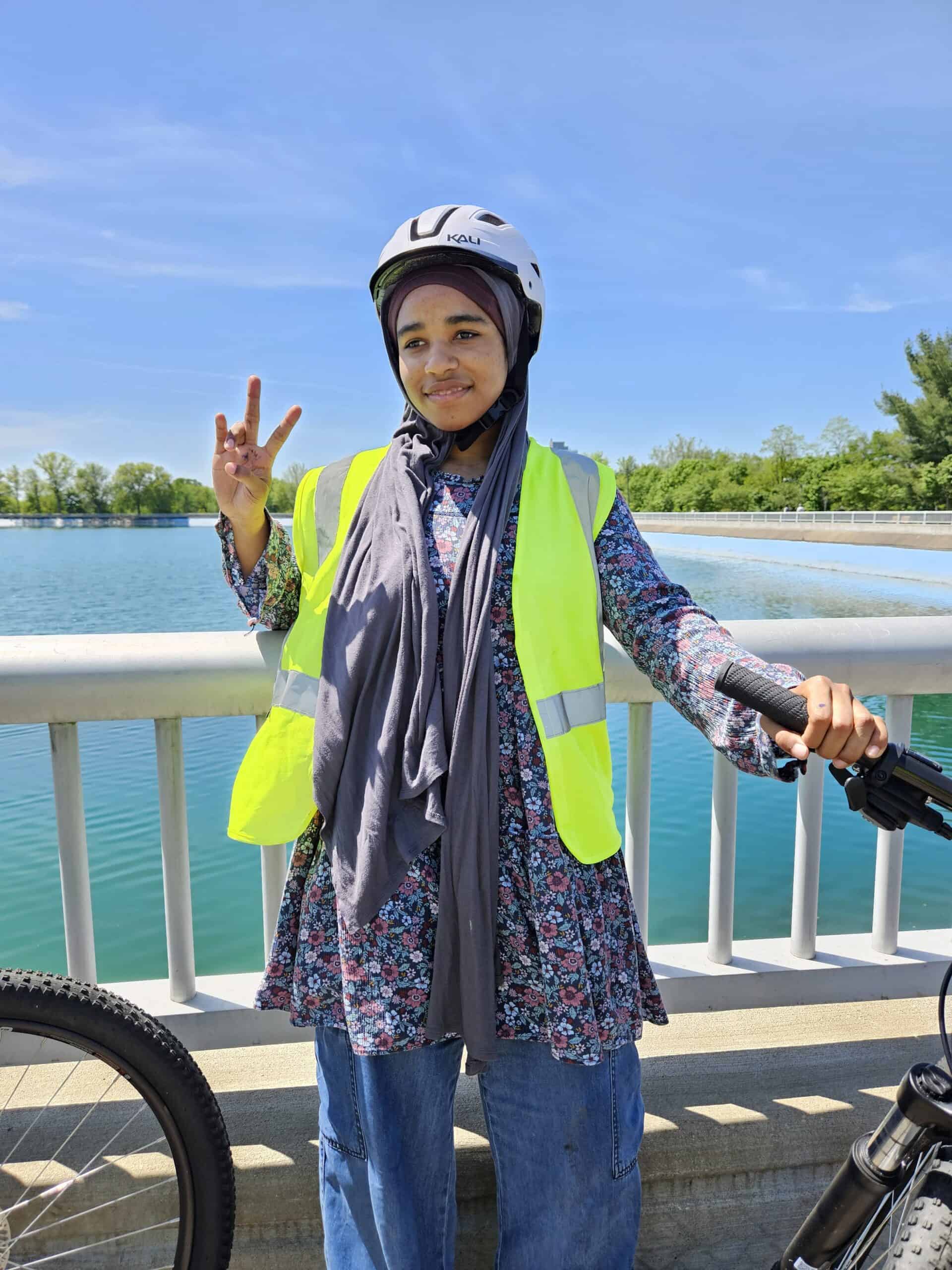 A female high-school student wearing a bike helmet and safety vest gives a "peace-sign" with one hand and holds the handlebars of her bike with the other. She is standing in front of a large reservoir in Highland Park.