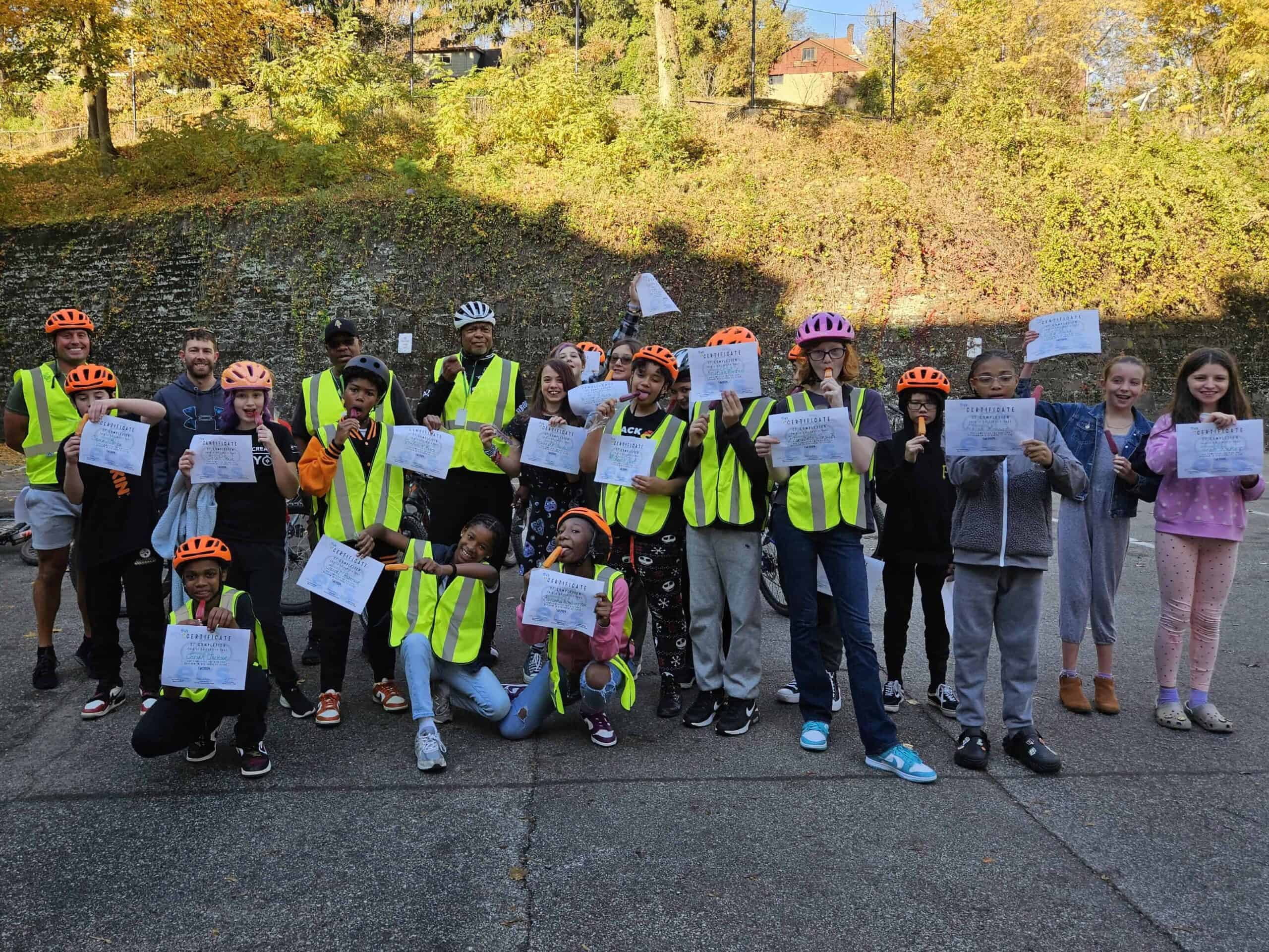 A group of roughly twenty middle school students wearing safety vests and helmets pose with paper certificates of completion while eating popsicles. 