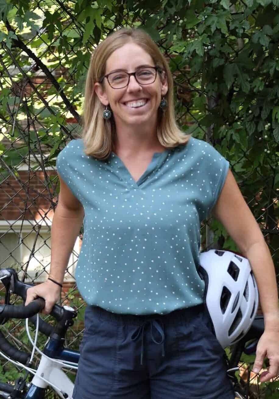 A woman with glasses and blonde hair smiles as she poses with her bicycle and helmet. 