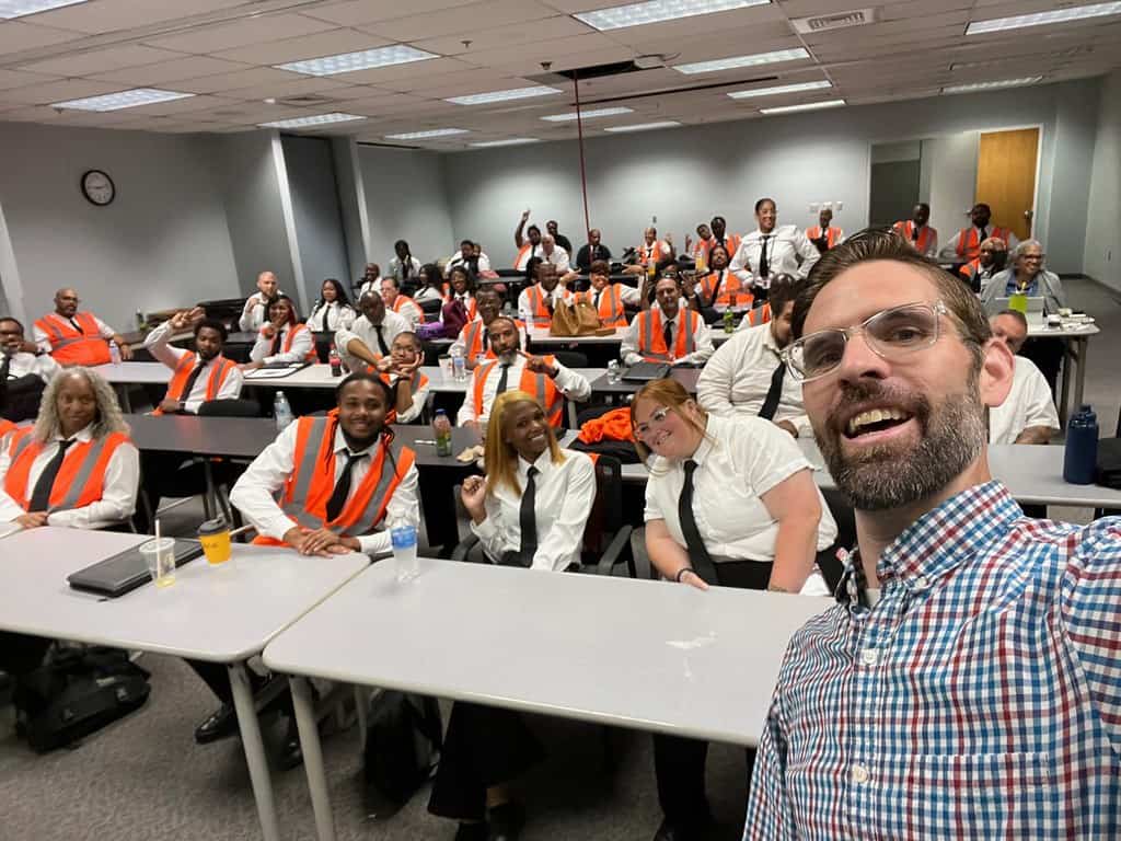 A middle-aged white man with glasses and a checkered shirt takes a selfie in a large classromm in front of fifty bus driver trainees. Trainees are wearing shirts and ties as well as orange safety vests. 