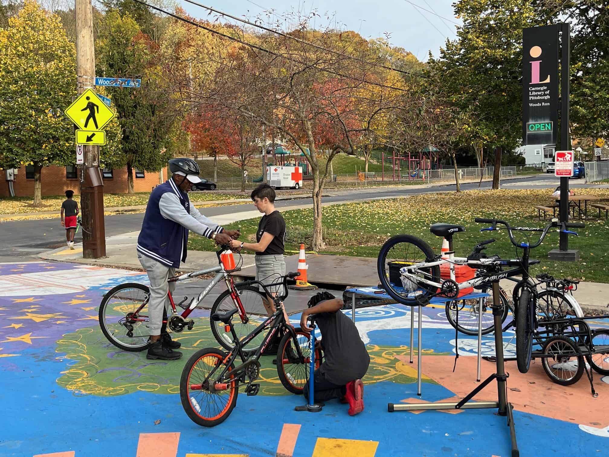 Pictured is a group of people standing on a street mural in front of a sidewalk surrounded by bikes and bike repair stands. A woman with short hair helps an older man who is straddling a mountain bike. In the foreground a teenage boy pumps up the tires of his bmx bike. 