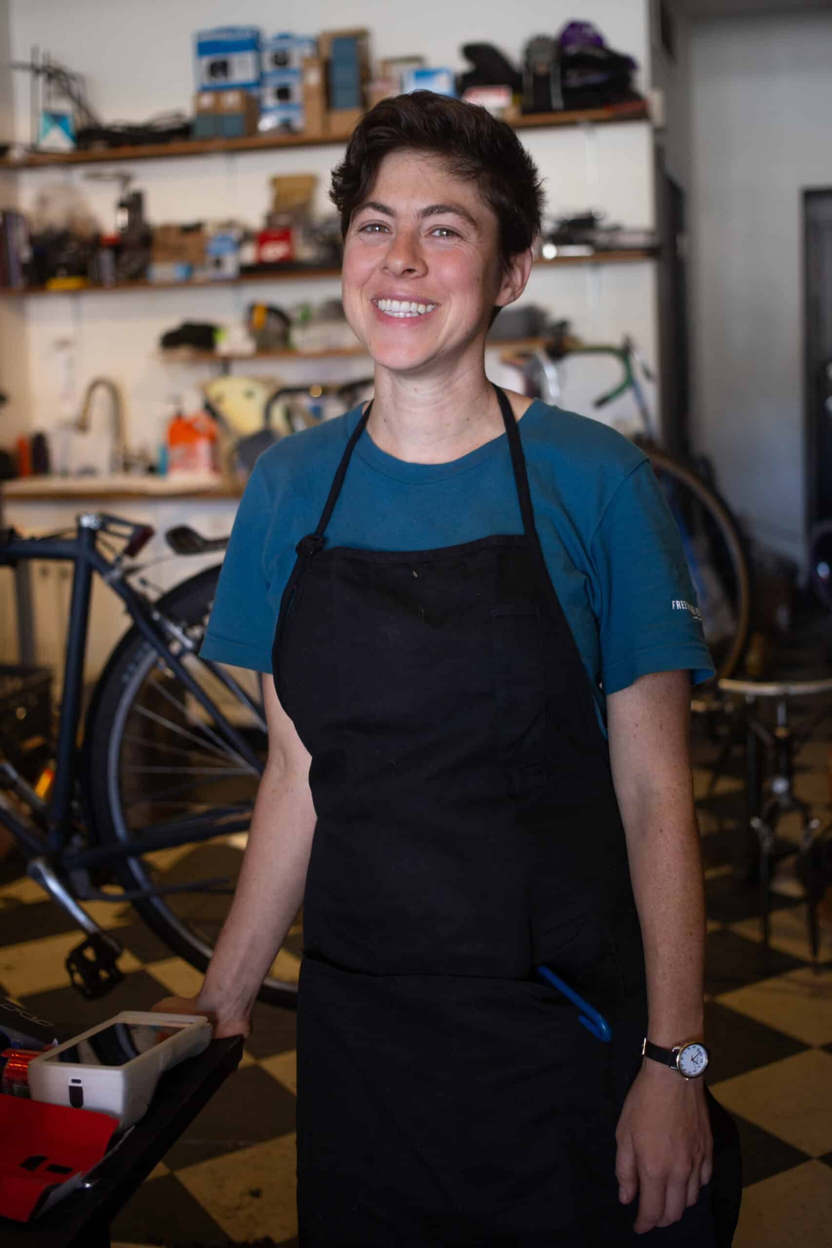 A woman with short hair wearing a shop apron smiles. Behind her are bicycles and racks of spare parts.