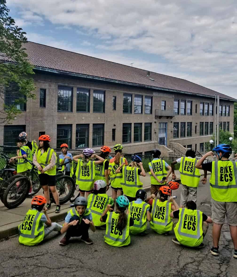 A group pof roughly twenty middle school students wearing safety vests that say "Bike ECS" and bike helmets sit in front of the ECS Middle School building after a bike ride. 