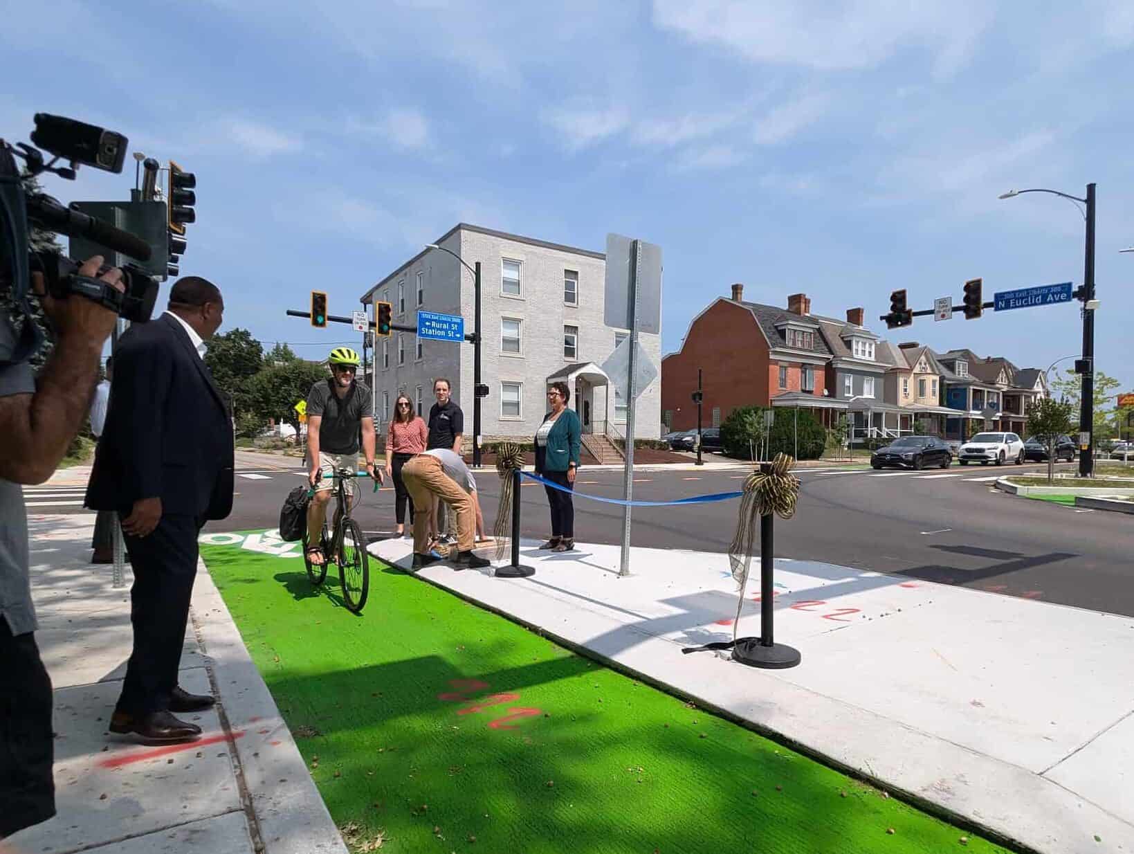 Image is of a green bike lane along N Euclid Ave. A bicyclists is riding down the lane while Mayor Gainey and a news camera operator is standing along the sidewalk