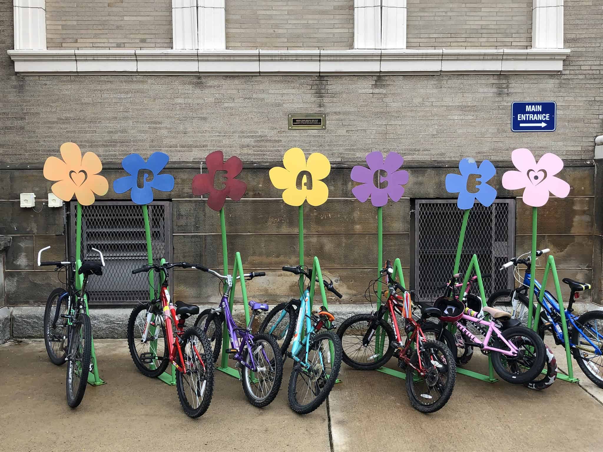 A green bike rack designed to look like the stems of flowers has bikes parked on it. The blossoms of the flowers spell out the word"peace." 