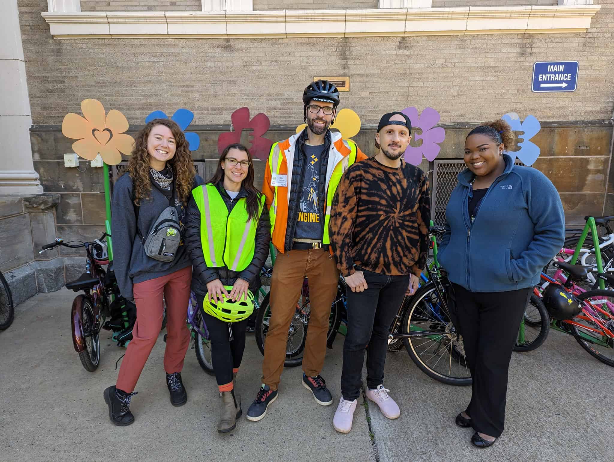 From left to right: a fair-skinned woman with curly hair is smiling, another fair-skinned woman with long brown hair and glasses smiles while holding a bike helmet. Next to her, a tall fair-skinned man with a bike helmet and glasses smiles, next to him another man with a backwards baseball cap and tie-dyed shirt smiles, to their right a dark-skinned woman with curly hair pulled back smiles. They are all standing in front of a bike rack filled with kids bikes. 