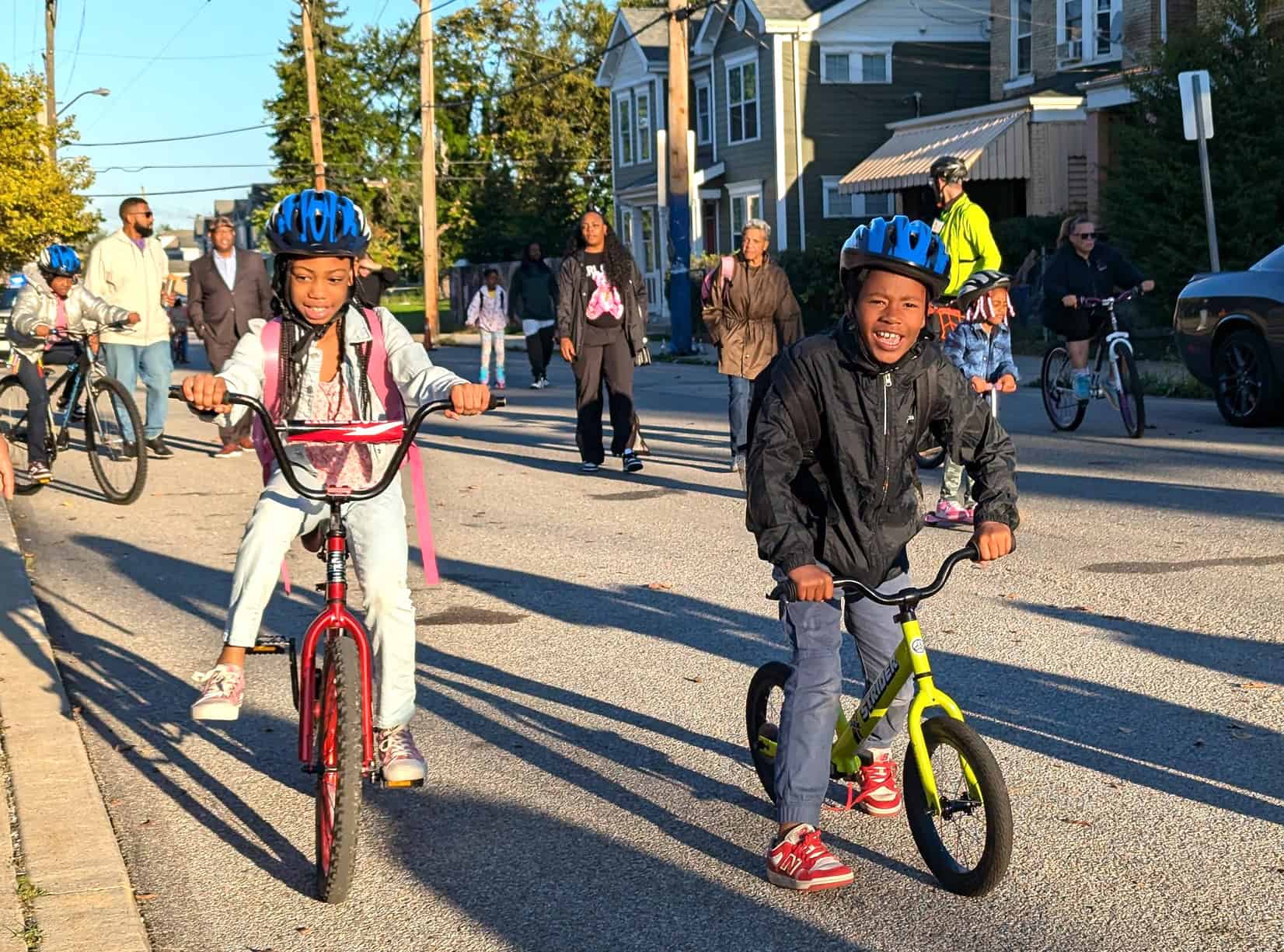 In the foreground are two smiling black children wearing bike helmets and riding bicycles. Behind them are other children riding bikes as well as some adults biking and walking. 