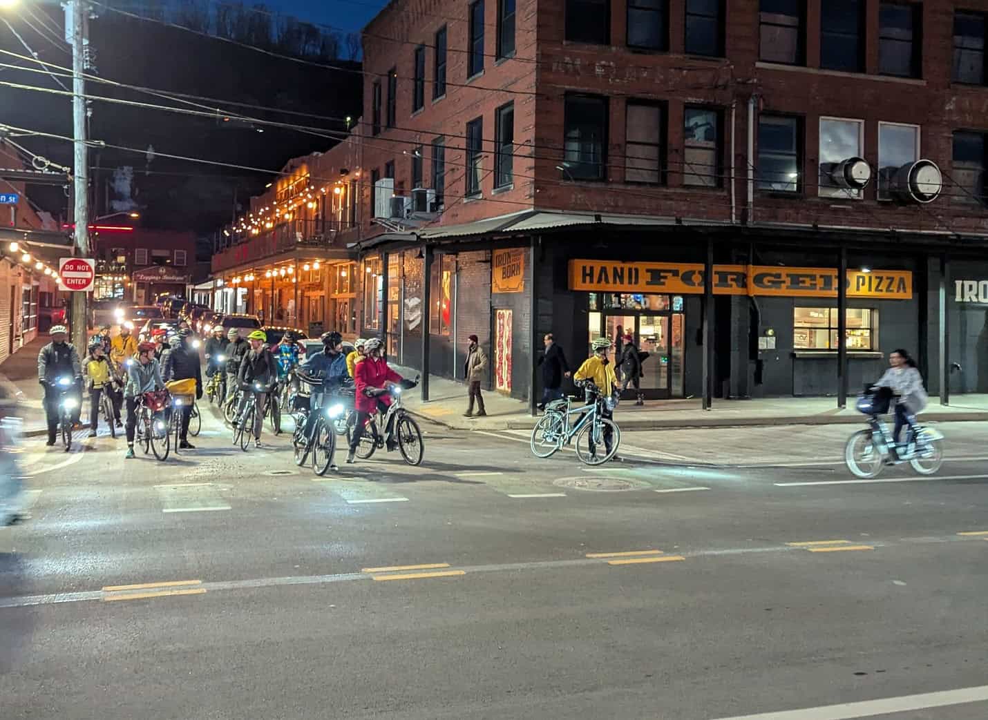 Cyclists at an intersection with their bikes waiting to proceed. Iron Born Hand Forged Pizza is in the background.