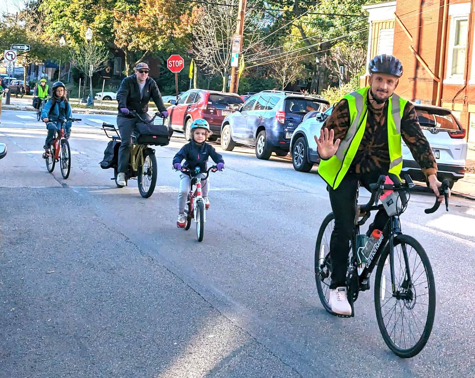 A fair-skinned adult man wearing a bright safety vest waves while riding a bicylce. Behind him are children and parents riding bikes on a city street. 