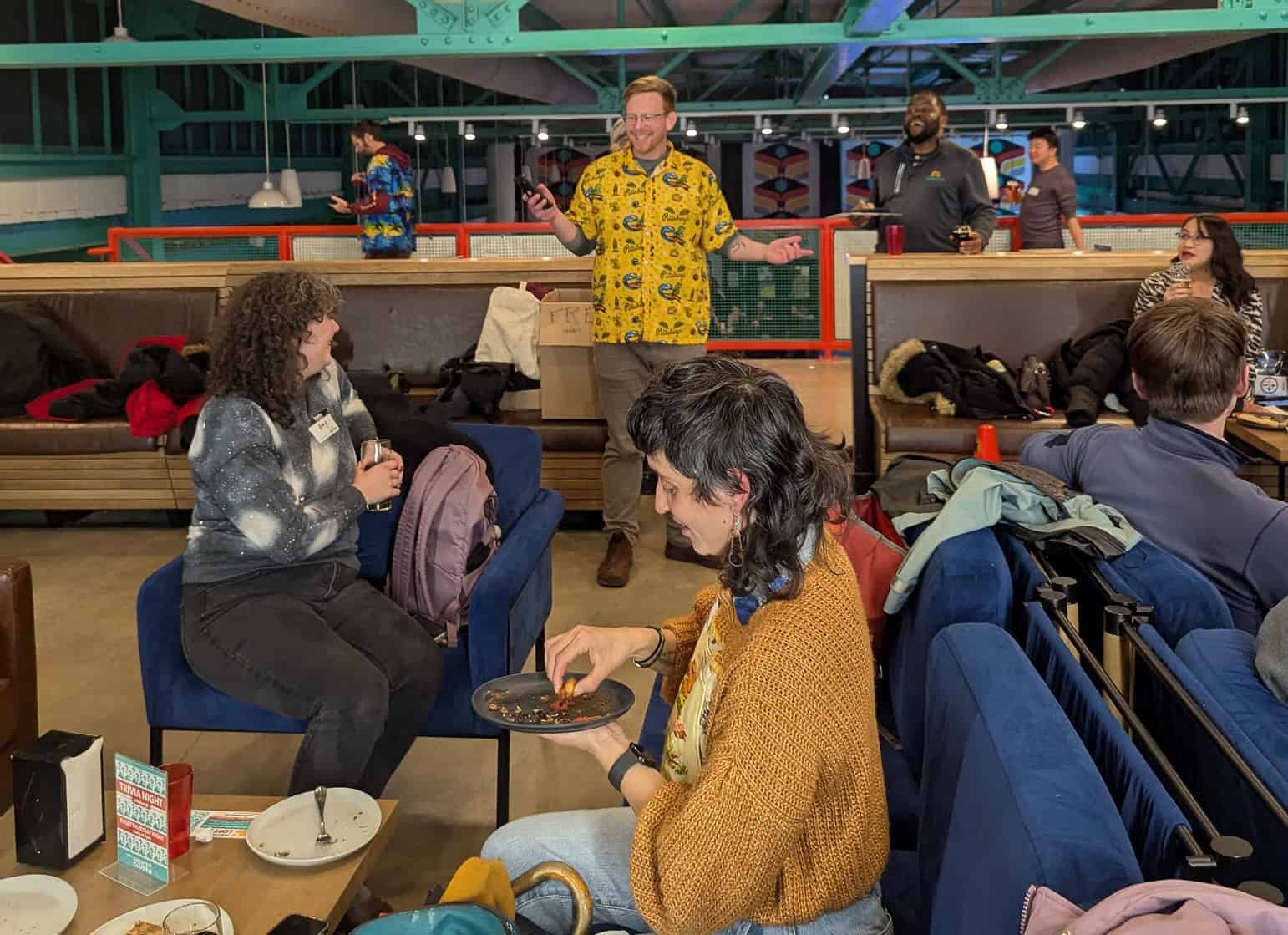 A person with red hair and a yellow tropical shirt laughs as he speaks with an audience of smiling advocates.