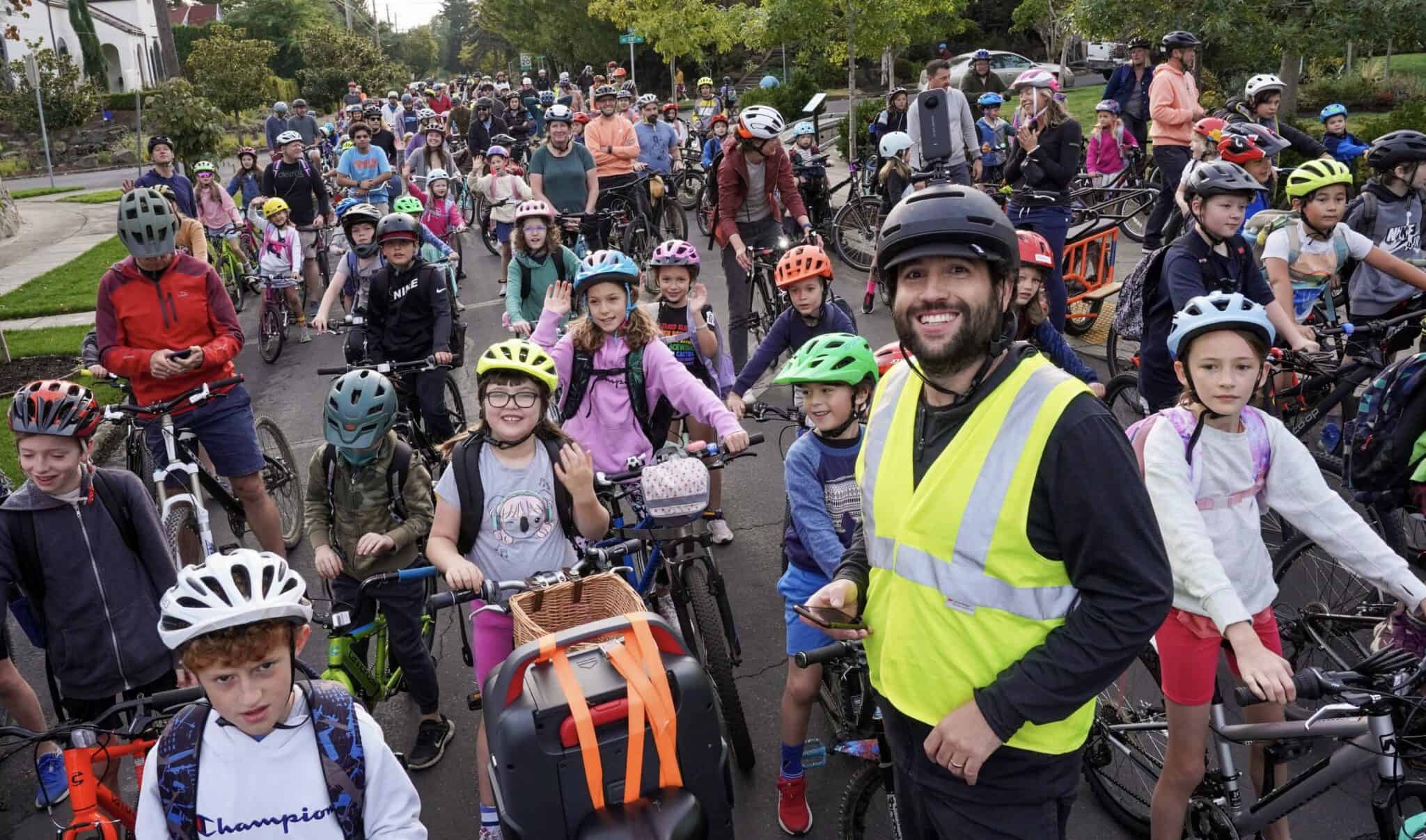 Sam Balto, foreground, is a smiling man with a dark beard, black helmet, and bright yellow safety vest. Behind him is over fifty children and adults astride bicycles on a road getting ready to bike to school. 