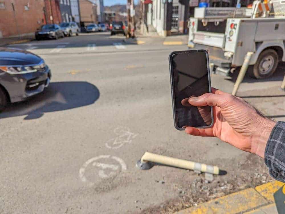 Image is of a person holding a phone using it snap a picture of a broken bollard and reporting it to 311.