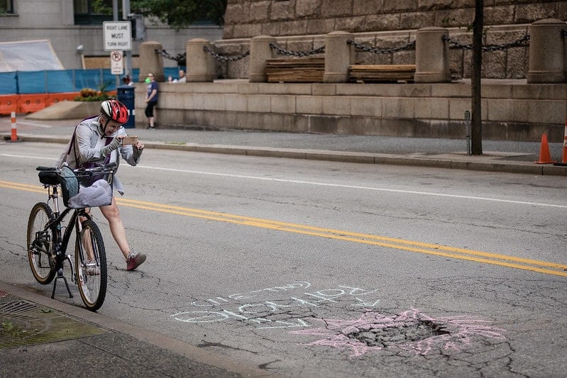 Photo of a person wearing a helmet taking a picture of a pothole with their bike off to the side. The pothole has street chalk around it to make it more visible.