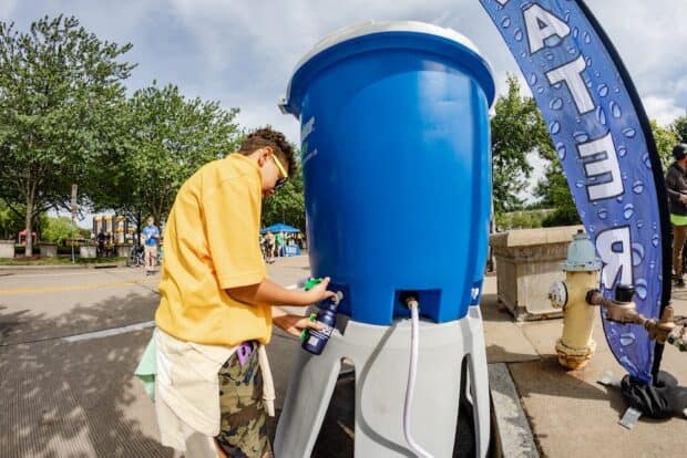 A person fills up their water bottle at a large water jug station next to a fire hydrant and a big blue sign that says WATER on it