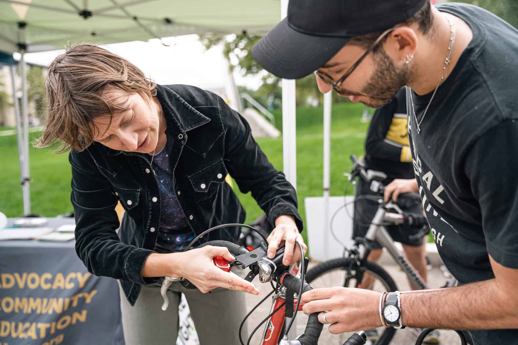 A woman demonstrates how to turn on a bike light that she has just mounted on the handlebars of a man's bike. Behind them is the BikePGH tent and a tablecloth that says "advocacy, community, education."