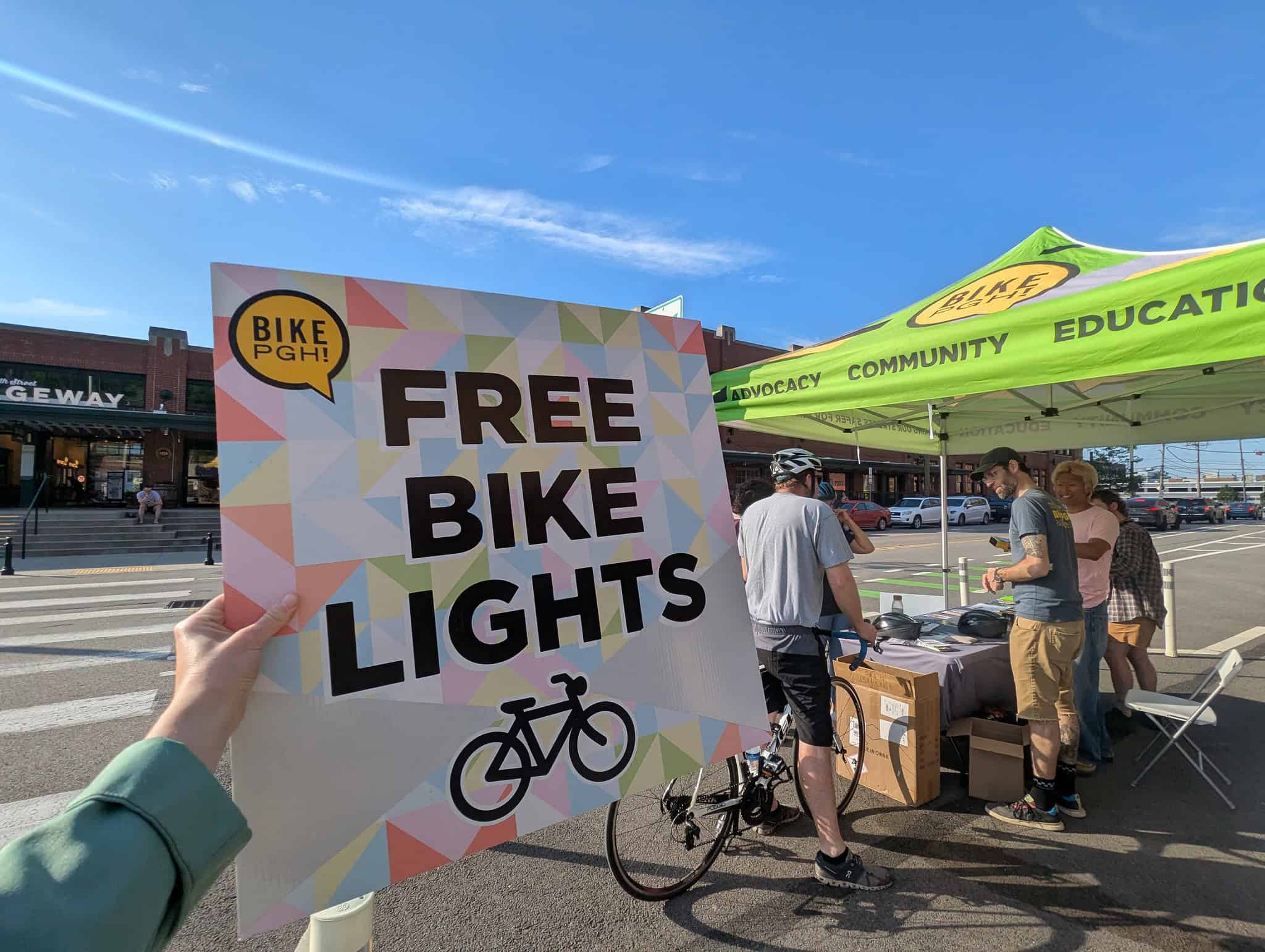 A hand holds a bright sign that says Free Bike Lights with a silhouette of a bicycle and the BikePGH logo. Behind the picture is a tent with BikePGH staff giving out supplies to a passing bike rider.