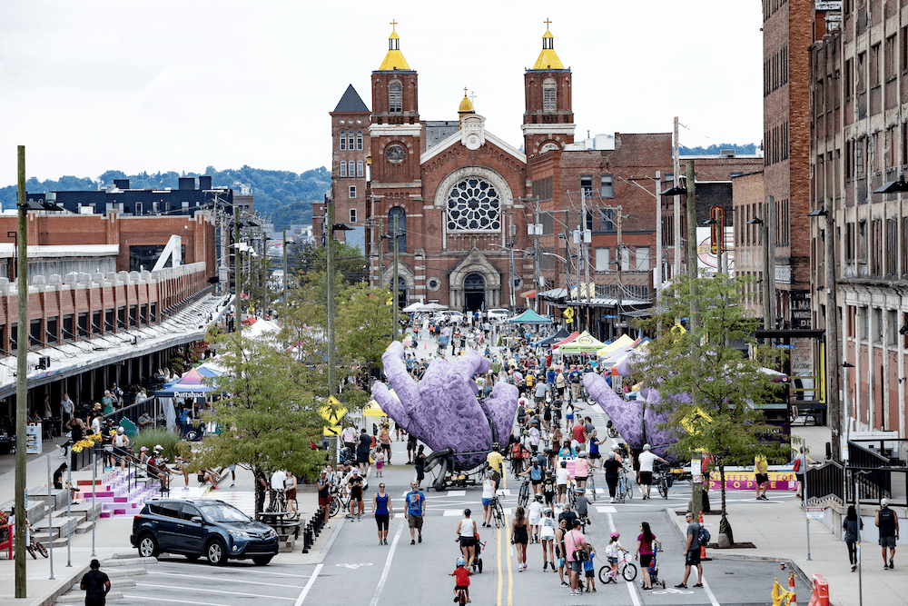 A zoomed out view of the Strip District filled with people walking, biking, and rolling on Smallman Street, with Squonk's big purple hands in the center, historic sights and hundreds of unique businesses and vendors tents.