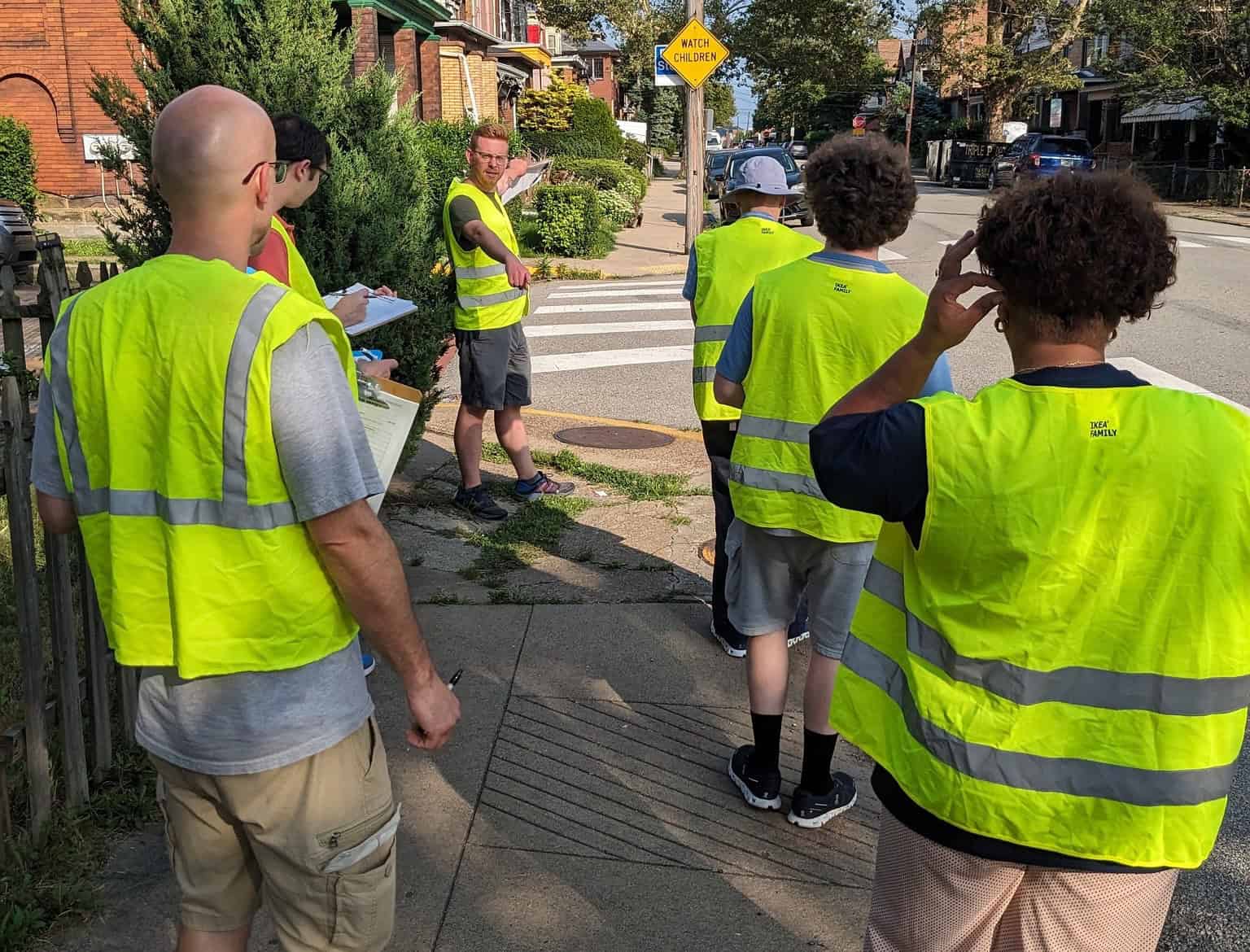 People wearing hi-vis vests take notes while observing a broken sidewalk and missing ADA ramp.