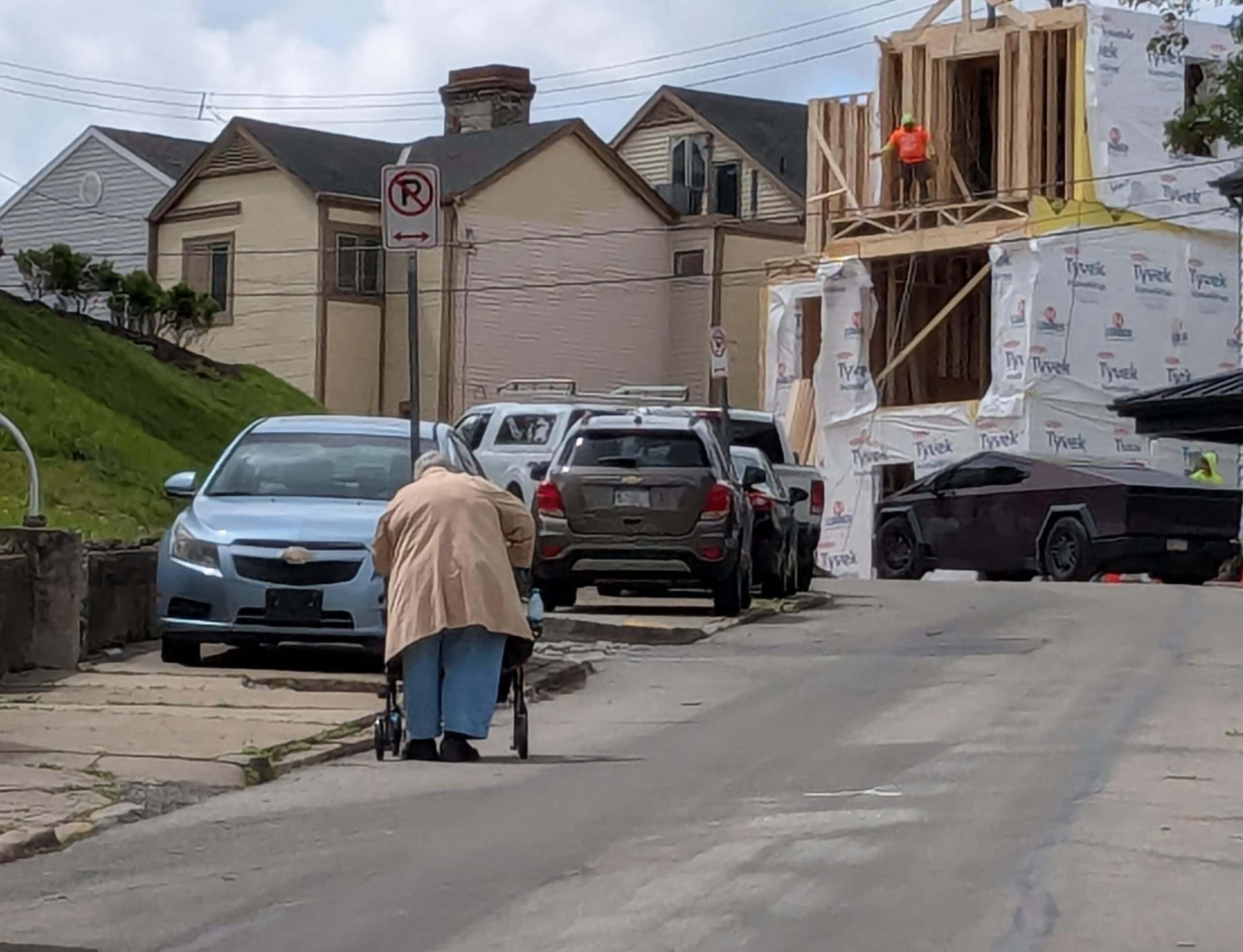 A person using a walker ascends a steep street in the roadway as cars are parked on the deteriorating sidewalk. An unreasonably large cybertruck adds insult to injury.