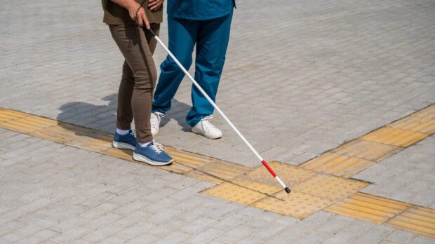 A waist down view of two people, one of which is using a white cane to feel the varying textures of yellow tactile pavers in the sidewalk.