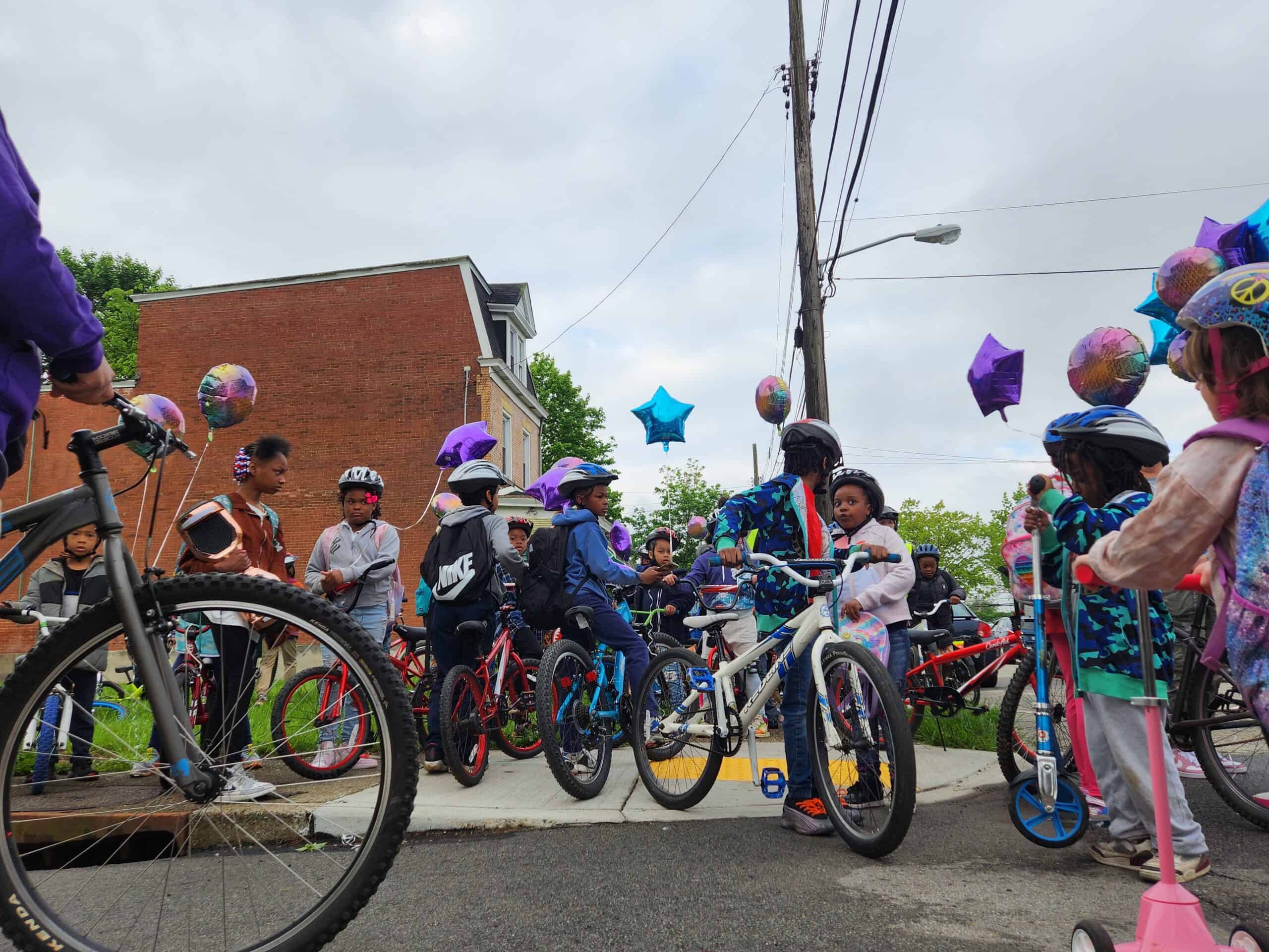 Several dozen children with helmets stand with with their bikes and scooters while holding balloons.