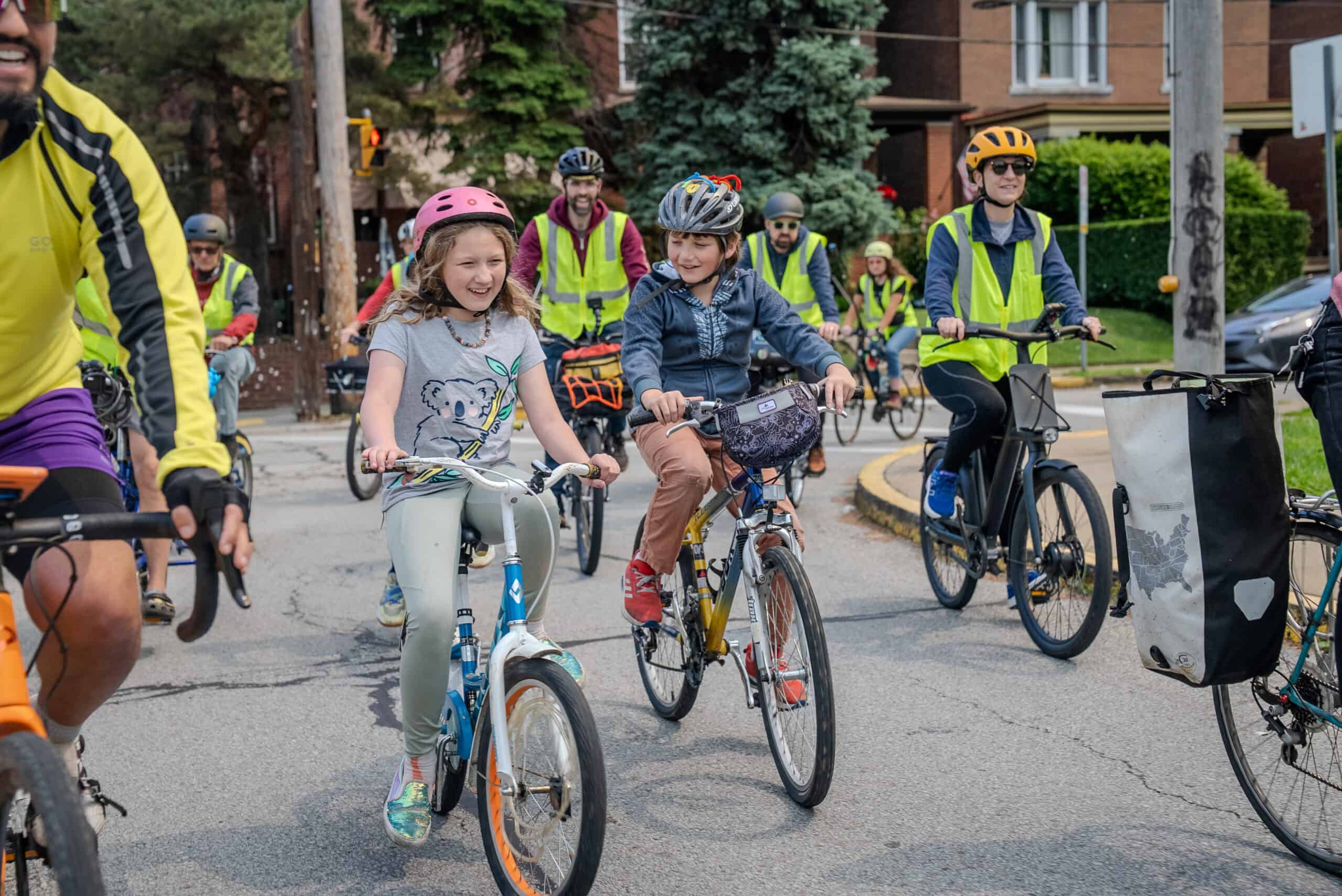 A boy and a girl riding to school are smiling. They are flanked by adult safety volunteers with neon yellow vests.