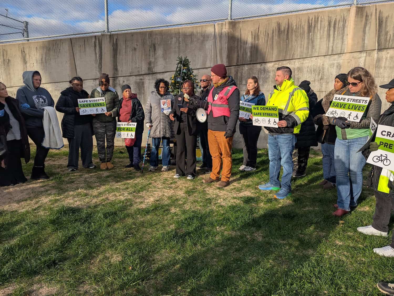 People gather around a memorial tree with names of victims of traffic violence. People are holding signs that say "safe streets for all" and "we demand safe streets".