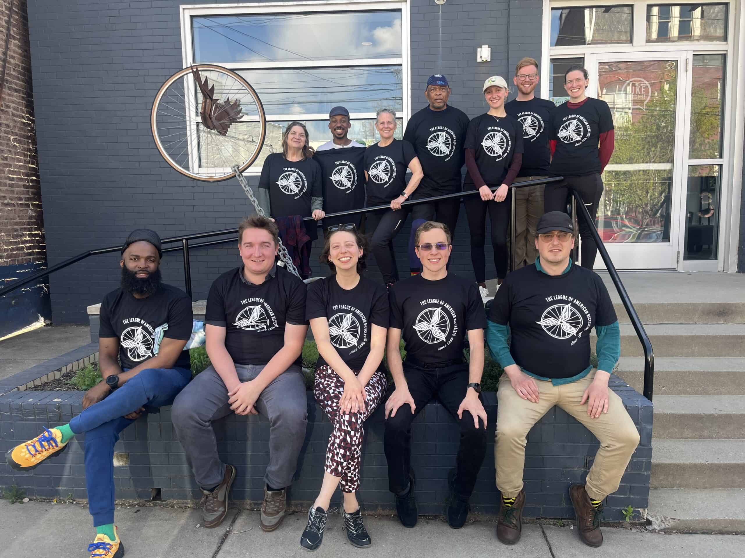 A group of 12 folks standing at the front step of the BikePGH office, all wearing matching black LCI shirts