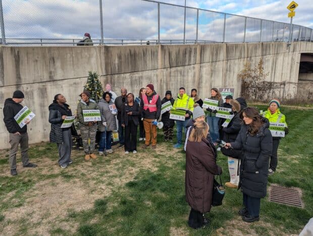 People gather around a memorial tree with names of victims of traffic violence. People are holding signs that say "safe streets for all" and "we demand safe streets". A reporter interviews someone.