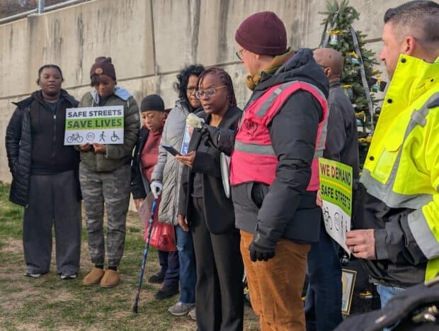 People gather around a memorial tree with names of victims of traffic violence. People are holding signs that say "safe streets for all" and "we demand safe streets".
