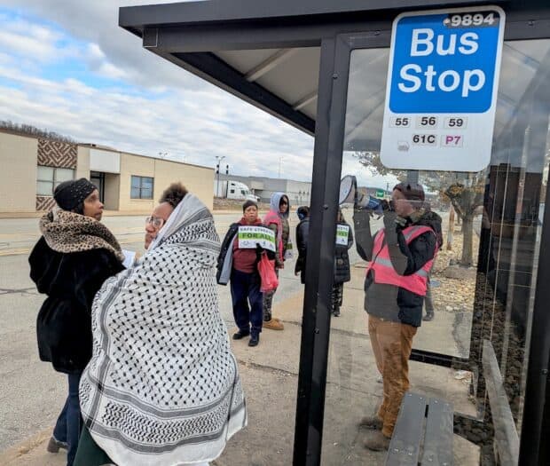 A man in a pink vest speaks through a megaphone at a bus stop.